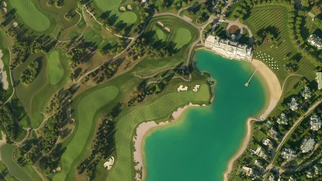 Aerial overhead view of a turquoise lake next to a golf resort with beautiful abstract patterns, sand traps, trees tennis courts, and a . Dolly shot with camera moving forward.