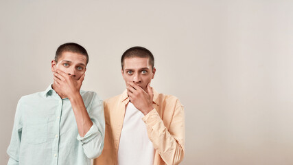 Two young scared twin brothers covering mouth with hand and looking at camera with shocked face expression while standing isolated over beige background