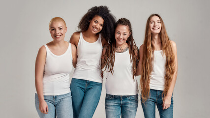 Portrait of four young diverse women wearing white shirts and denim jeans smiling at camera while...