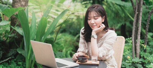 Young Asian woman working with laptop computer and drinking a cup of coffee in a garden. A woman with smiley face working from home. Social distancing concept. Panoramic banner portion
