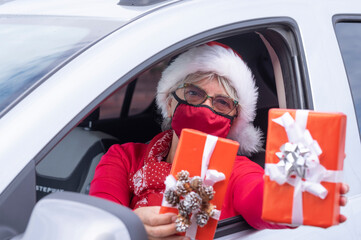 Coronavirus and Christmas time. A senior woman wearing a Santa hat and medical mask to avoid coronavirus infection drive the car for home delivery of Christmas gifts.