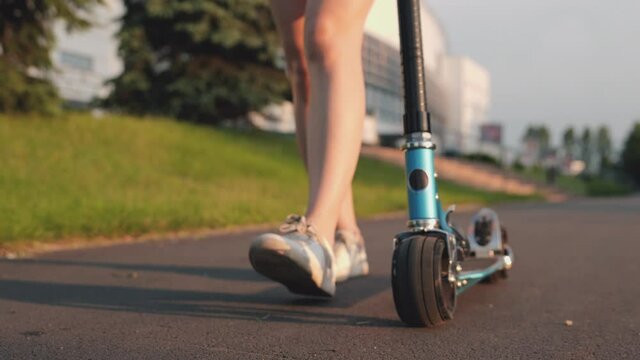Close-up foot or leg of young woman walking on sidewalk with blue scooter. Girl walking in park on sunset. Teenager spends time alone at sunlight. Concept of college life, skating, summertime.