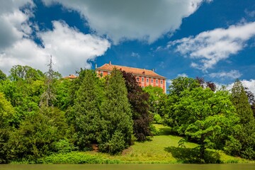Fototapeta na wymiar Opocno, Czech Republic - June 16 2020: View of the castle with red facade standing on a hill from a park with green trees and grass. Bright sunny summer day with blue sky and white clouds.