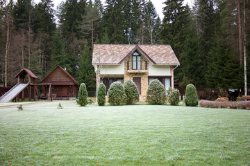 Country house with green lawn in background of forest on frosty autumn day. Natural landscape in winter. Beauty of nature concept.
