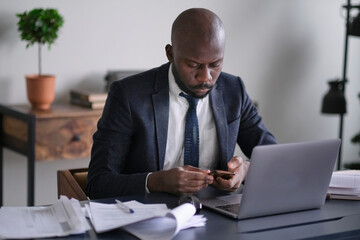 Young African executive uses a smartphone, reads bad news and working on a laptop while sitting at his desk in an office. soft focus