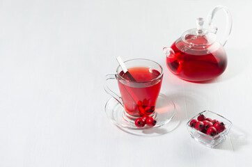 Red hawthorn tea in transparent teapot with cup, fresh berries in sunlight with shadow on white wood board, copy space.