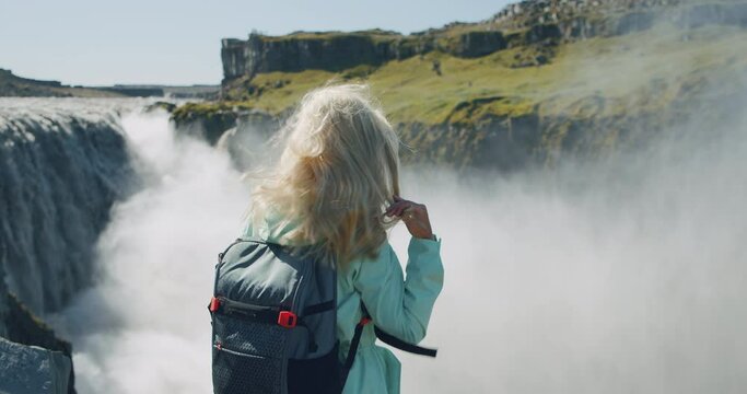 Iceland. Rear close up of blond hair woman standing at cliff edge with beautiful detifoss waterfall in background