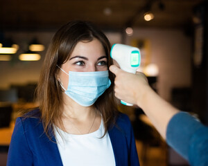 Doctor measures the body temperature of a young woman in a protective mask with an infrared...