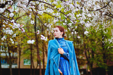 portrait of a redhaired young woman in a blue coat and retro dress in a rainy spring garden of apple trees. 