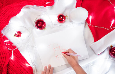 Beautiful creative Christmas background with pattern. The child draws Santa Claus with a red pen on a white sheet and red Christmas balls are scattered around and a garland lights up.