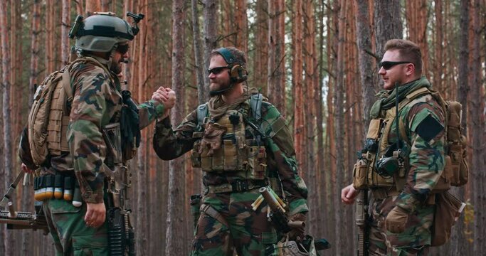 Fully equipped soldiers in camouflage uniform are enjoying the completed action in the forest. They get high fives, smile, and are happy