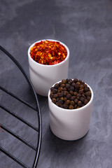 Whole black pepper and chilli flakes arranged in a white small bowl with grill beside with grey  textured background