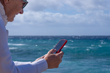 A senior woman white dressed enjoying beach vacation on a windy day reading a message in her smartphone.  Relaxed smiling active retiree