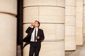 Middle aged buisnessman drinking takeaway coffee while leaning on column in street with cellphone in his hand