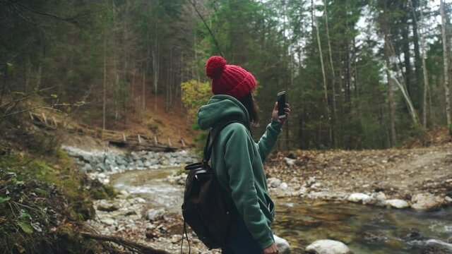 Woman in hike wear with backpack take panorama shots on smartphone or mobile phone while standing on shore of mountain river. Millennial using modern technology to making or taking photo for memory.