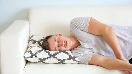 Tired young man with closed eyes in white t-shirt lies on soft couch with small designer pillow in light room at home close-up