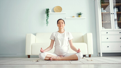 Smiling young pregnant lady in white meditates sitting in padmasana yoga position on floor against...