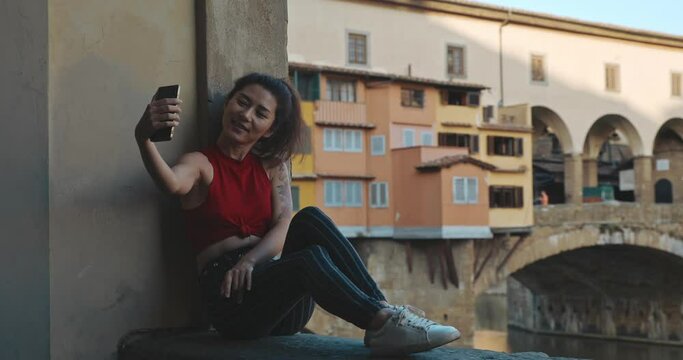 Portrait of young woman travelling to Florence during her summer vacation. Asian girl in red T-shirt and striped blue jeans sitting on Vecchio bridge and making selfie on her smartphone.