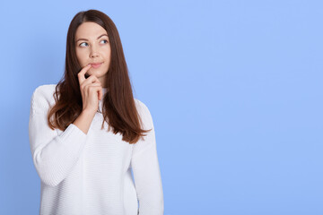 Young pensive girl with dark hair, keeps fore finger near corner of lips, looking aside while pondering on idea, has doubts, wears casual white shirt, models over blue background.