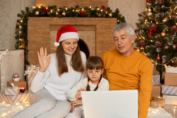 Fototapeta na wymiar Photo of grandad, mom and child making x-mas eve video call, meeting conference, waving arms, congratulating relatives, sitting indoors wearing santa claus cap and casual clothing.