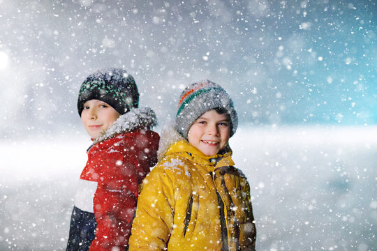 Two little kids boys of elementary class walking to school during snowfall on early dark morning. Happy children having fun and playing with first snow. Siblings ans friends in colorful winter clothes