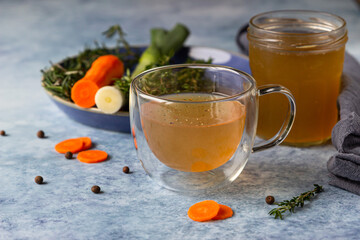 Homemade bone broth in glass mug and vegetables, blue concrete background. Collagen source for the body.