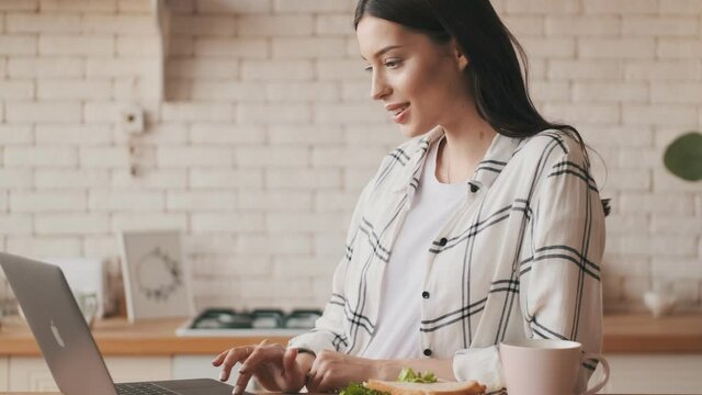 Happy pregnant woman using laptop computer at the kitchen while having a breakfast indoors at home