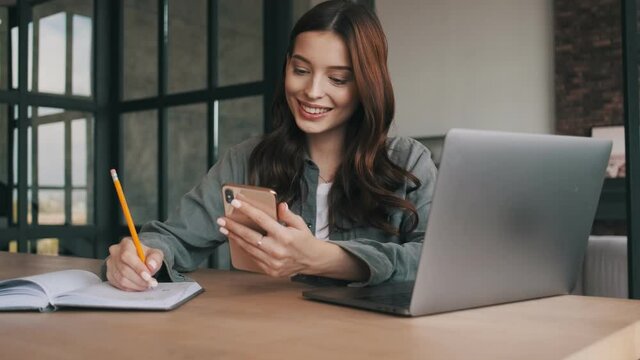 Happy young woman using mobile phone and laptop computer while writing notes indoors at home