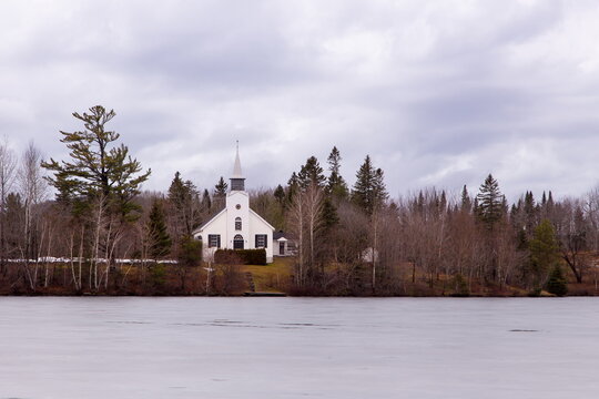 White catholic chapel surrounded by woods seen across a lake during a grey misty winter day, Lac-Beauport, Quebec, Canada