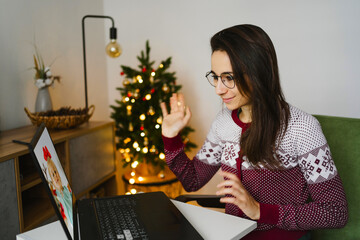  Young woman give gifts online on laptop sitting in room with christmas  festive decorations. Girl make a video call to relatives on holiday near the Christmas tree at home. Christmas party online..