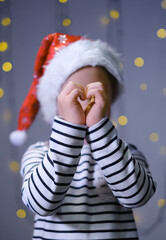 A small Caucasian child in a Christmas Santa hat made a heart with his hands at night in a room decorated with garlands. New year's portrait of a child with a heart on a blue background with lights