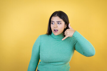Young caucasian woman over isolated yellow background confused doing phone gesture with hand and fingers like talking on the telephone