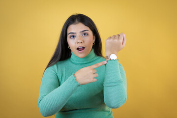 Obraz premium Young caucasian woman over isolated yellow background surprised and pointing her watch because it's late
