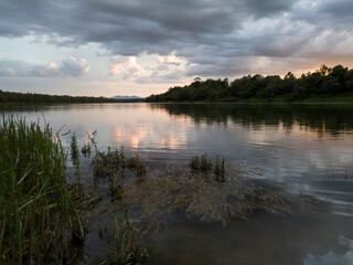 Landscape with river, clouds, tall water grass and mountain in distance at sunset. Peaceful riverine scene during beautiful cloudy evening.