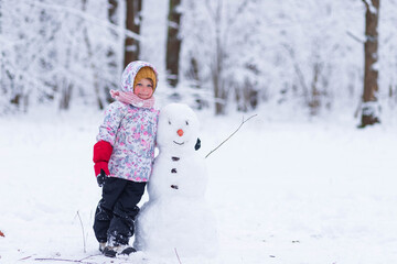 Happy child plays with a snowman on a winter walk in nature in the forest. Funny happy little girl on a walk in the winter outdoors