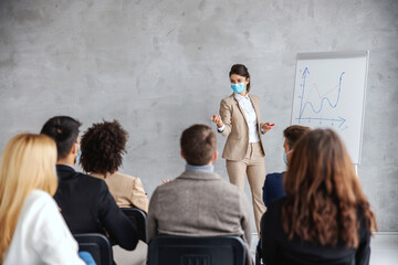 Young businesswoman with face mask standing in front of audience an having presentation of monthly...