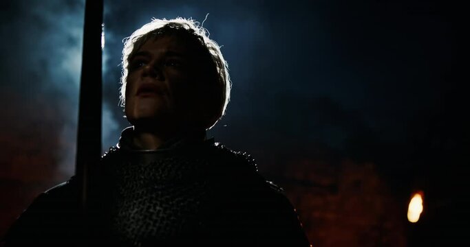 Silhouette of Joan of Arc with medieval sword looking up and praying against smoke and fire on dark battlefield. Female warrior with sword on battlefield
