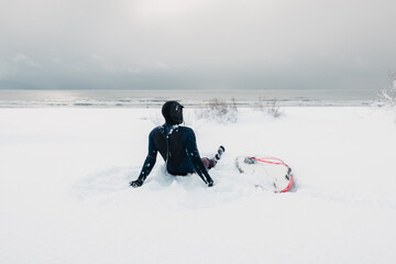 Cold winter and surfer sitting on snow beach with surfboard. Winter with surfer in wetsuit.