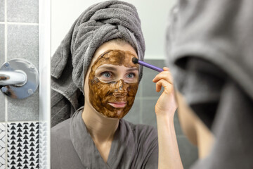 woman applying mud facial mask with brush in front of mirror in bathroom at home. skin care beauty treatment
