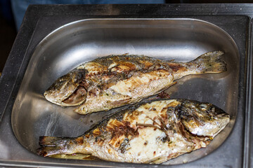 Two freshly cooked fried fish are in a metal container. Delicious food cooked over an open fire, which is offered at a street food fair, event, festival.