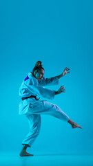 Fototapeta na wymiar Active training. Professional female judoist in white judo kimono practicing and training isolated on blue neoned studio background. Grace of motion and action. Healthy lifestyle, sport concept