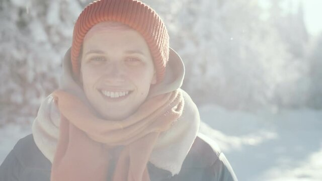 Portrait chest-up shot of young pretty woman in outerwear, hat and scarf looking at camera and happily smiling while standing in forest on sunny winter day