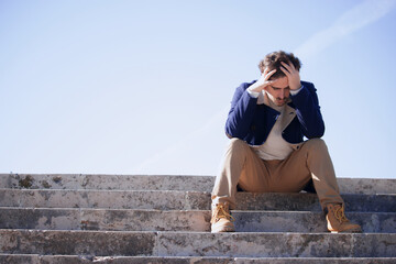Fototapeta premium A young man aged 24-30 sitting in the street frustrated after being fired from his last job. Disgruntled man loses his office job.