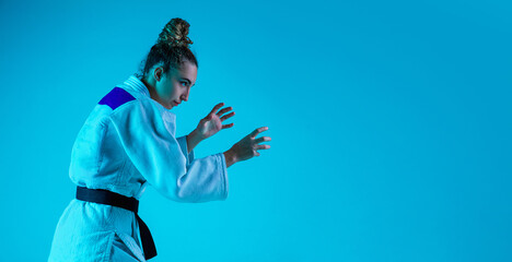 View from side. Professional female judoist in white judo kimono practicing and training isolated on blue neon background. Grace of motion and action. Healthy lifestyle, sport and movement concept.