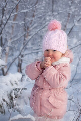 little girl todller pink down jacket pink knitted hat walks snowy Park Christmas day.The baby cold warms her hands.