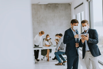Two young business men with facial protective masks discussing  with paper plan in the office
