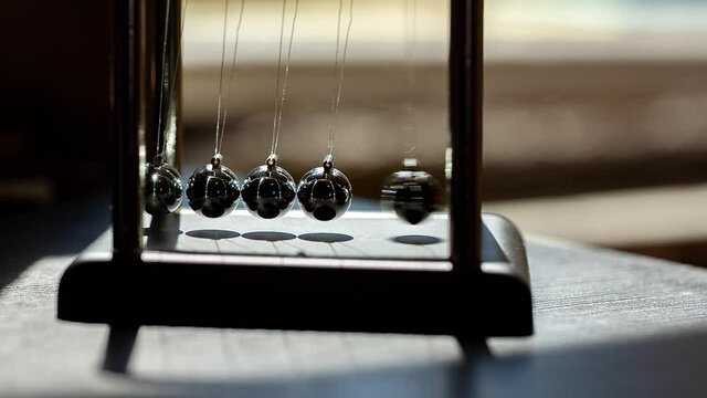 Hand putting a simple Newtons cradle in motion in sunlight, macro, closeup. Metal balls swinging and slowing down. Moving something, inertia concept, science, fun physics lesson toy, real time