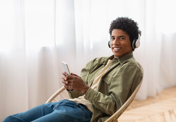 Handsome black adolescent in headset picking music playlist on mobile phone at home