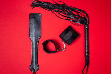 BDSM accessories on a red background. A leather paddle, padded handcuffs, and a whip lie next to each other. Sex accessories