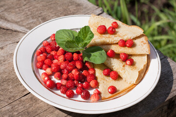 Red ripe wild strawberry with thin homemade pancakes on a white plate with a green fresh mint leaf.